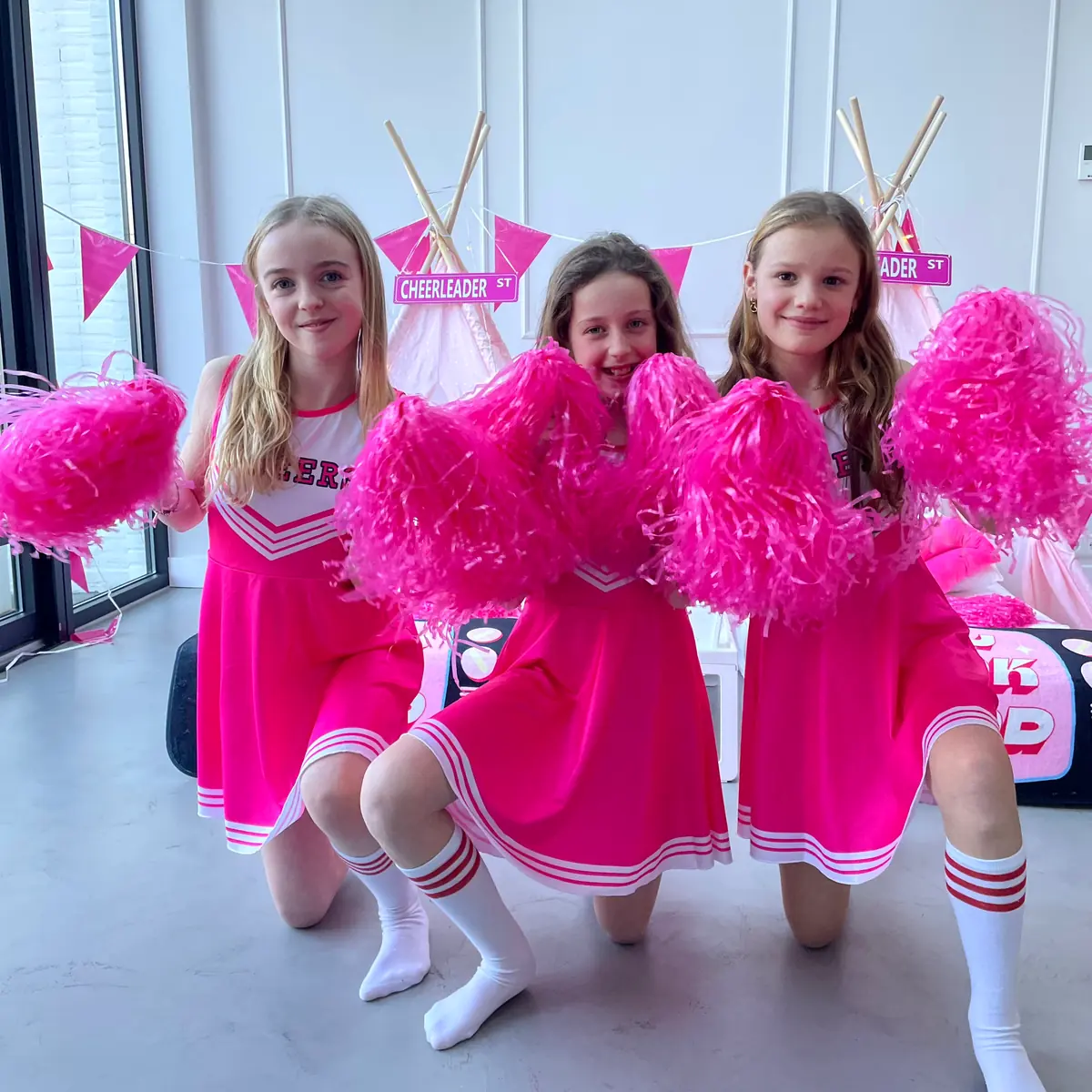 Drie meiden in roze cheerleaderjurkjes met pompons voor hun tipi’s